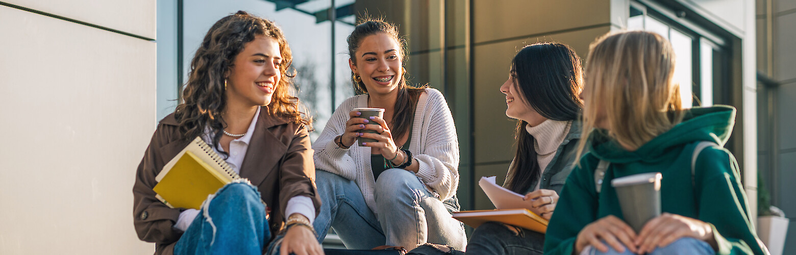 Studentinnen sitzen entspannt auf den Treppen vor der Hochschule, unterhalten sich und lernen gemeinsam – Campusleben, Studium und Studentenalltag in Deutschland