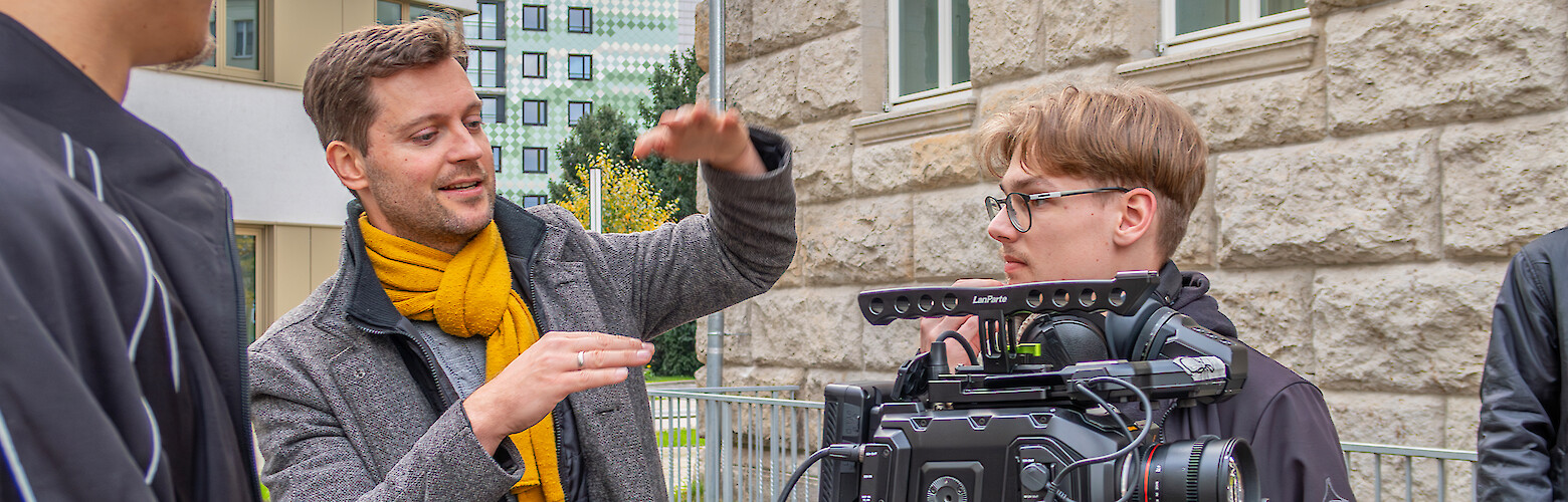 Dozent erklärt Studierenden den Kameraeinsatz während eines Praxisprojekts an der Fachhochschule Dresden. Szene findet im Freien vor dem Hochschulgebäude statt. Der Fokus liegt auf filmischer Arbeit und praktischer Anwendung im Design-Studiengang.