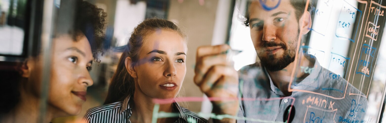 Zwei Frauen und ein Mann skizzieren Business Ideen an einer Glasscheibe.