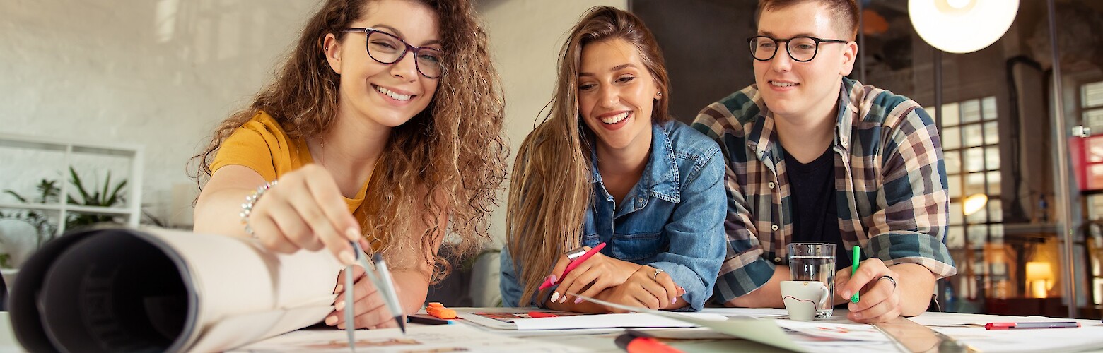 Zwei Frauen und ein Mann arbeiten am Schreibtisch an Skizzen mit Zirkel und Stiften.