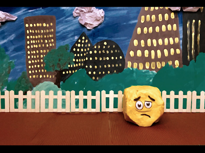Handgemachte Stop-Motion-Szene mit traurig dreinblickendem gelbem Knetball vor weißem Holzzaun; im Hintergrund gemalte Großstadt-Silhouette mit Hochhäusern, Bäumen und Wolken bei Nacht